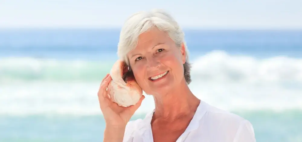Senior woman holding seashell to her ear at beach, smiling.